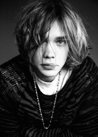 Portrait of a young man with tousled medium-length hair wearing a patterned knit sweater and a long chain necklace against a dark background.
