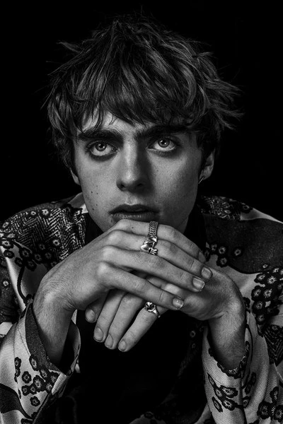 Black and white portrait of a young man with tousled hair and floral patterned shirt, looking at the camera with hands under chin.