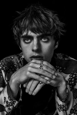 Black and white portrait of a young man with tousled hair and floral patterned shirt, looking at the camera with hands under chin.