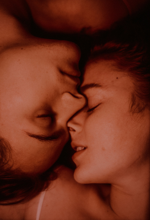 Close-up of a couple lying down with their eyes closed, face-to-face, noses touching, lips slightly parted, under warm-toned lighting.
