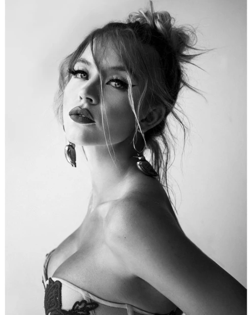 Black and white portrait of a fair-skinned woman with dark red lips, dangling bird earrings, and her blonde hair up in a messy bun.