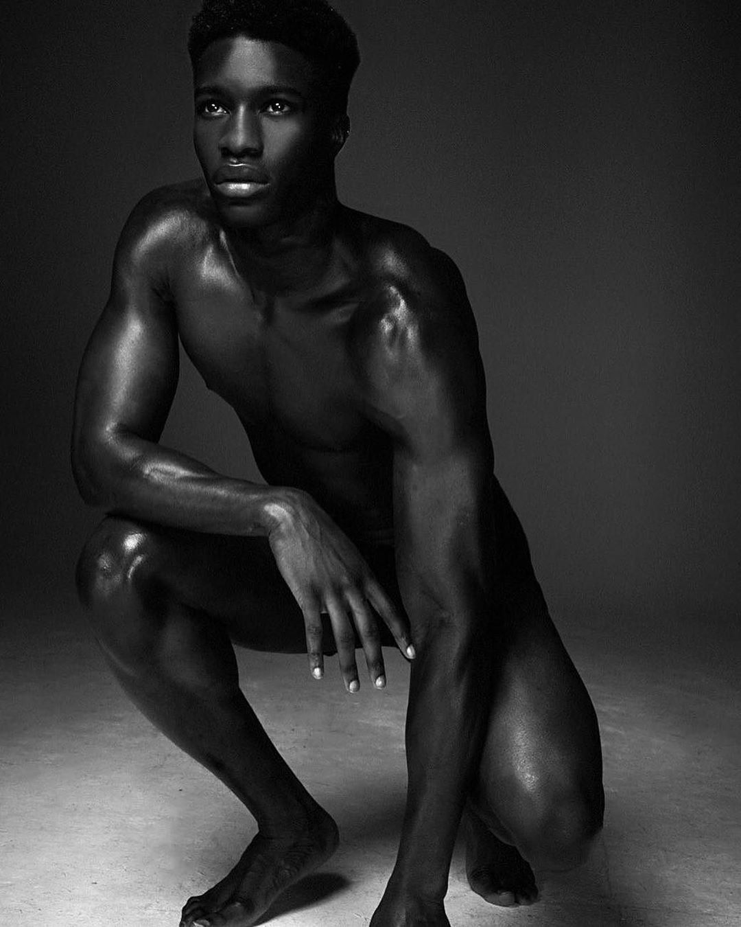 Striking black and white portrait of a muscular Black male model squatting in a photo studio, showcasing physique and posture.