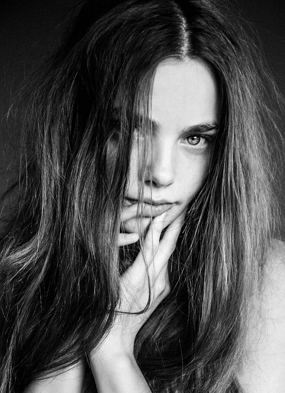 Black and white portrait of a young adult female with long, wavy hair partially covering her face, resting her chin on her hand.