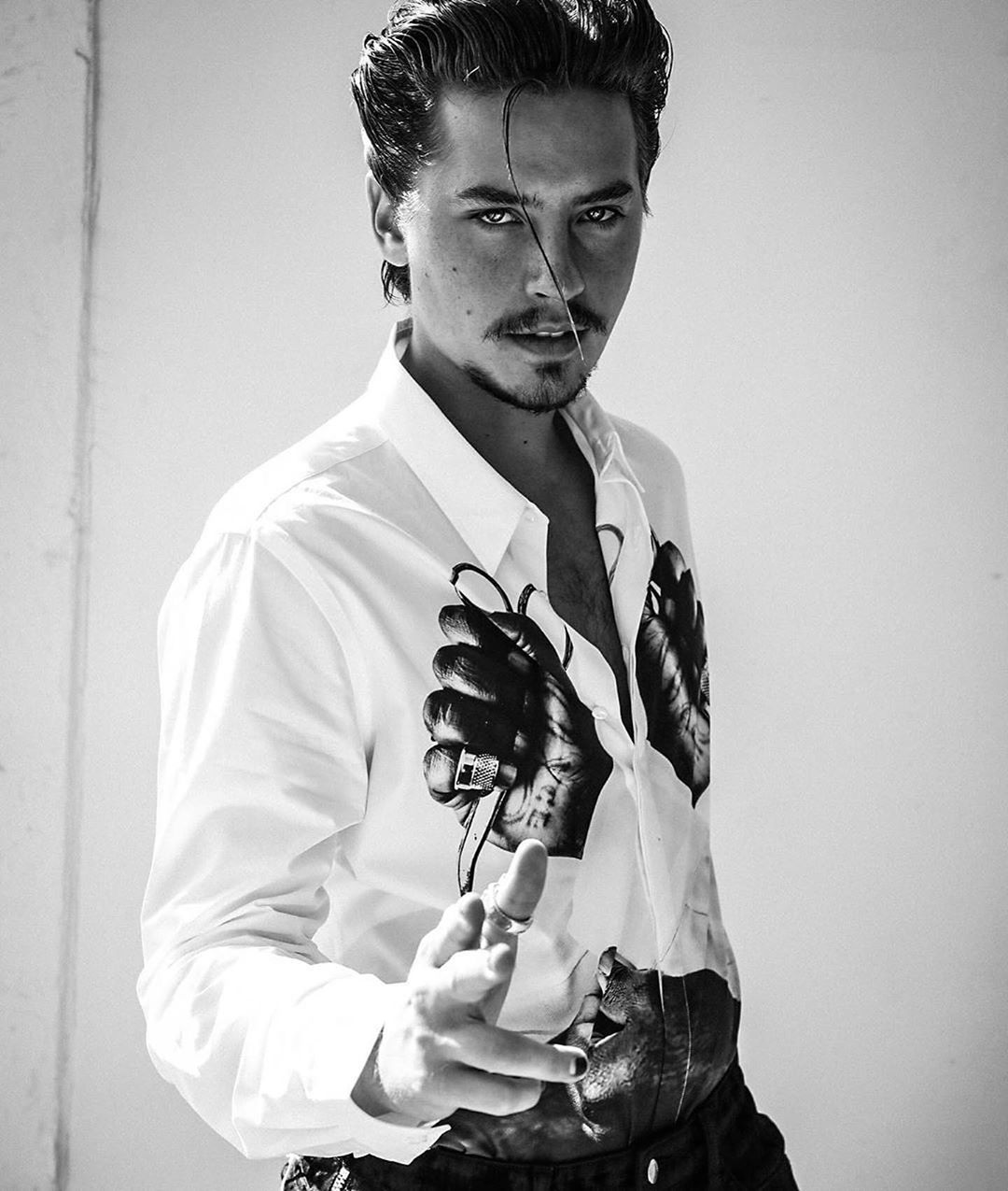 Black-and-white portrait of an adult male with styled hair, wearing a white shirt with black graphic prints, posing with outstretched hand.