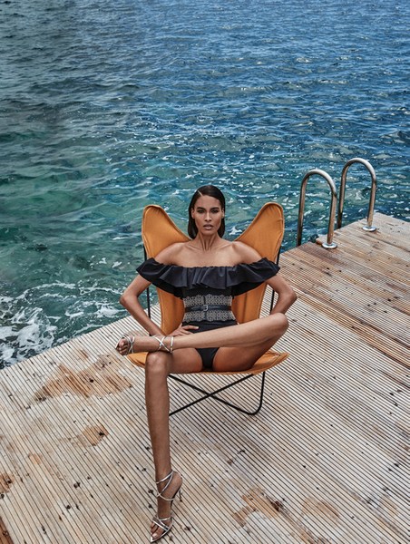 A tanned woman in a black ruffled swimsuit poses in a yellow chair on a wooden dock, set against the backdrop of a blue ocean.