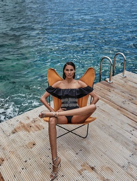 A tanned woman in a black ruffled swimsuit poses in a yellow chair on a wooden dock, set against the backdrop of a blue ocean.