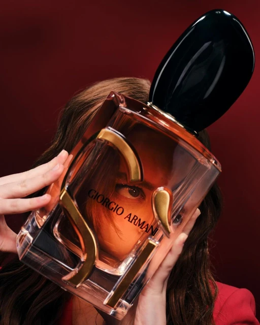 A woman holds a Giorgio Armani perfume bottle, partially obscuring her face, against a dark red backdrop in a beauty shot.