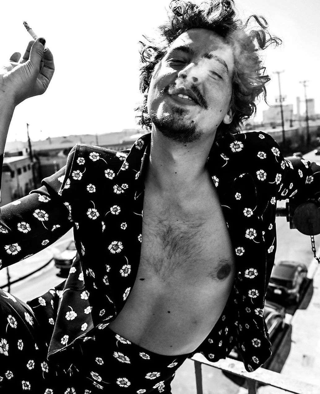 Black and white portrait of a man with curly hair and a floral-patterned suit smoking a cigarette on a city rooftop.