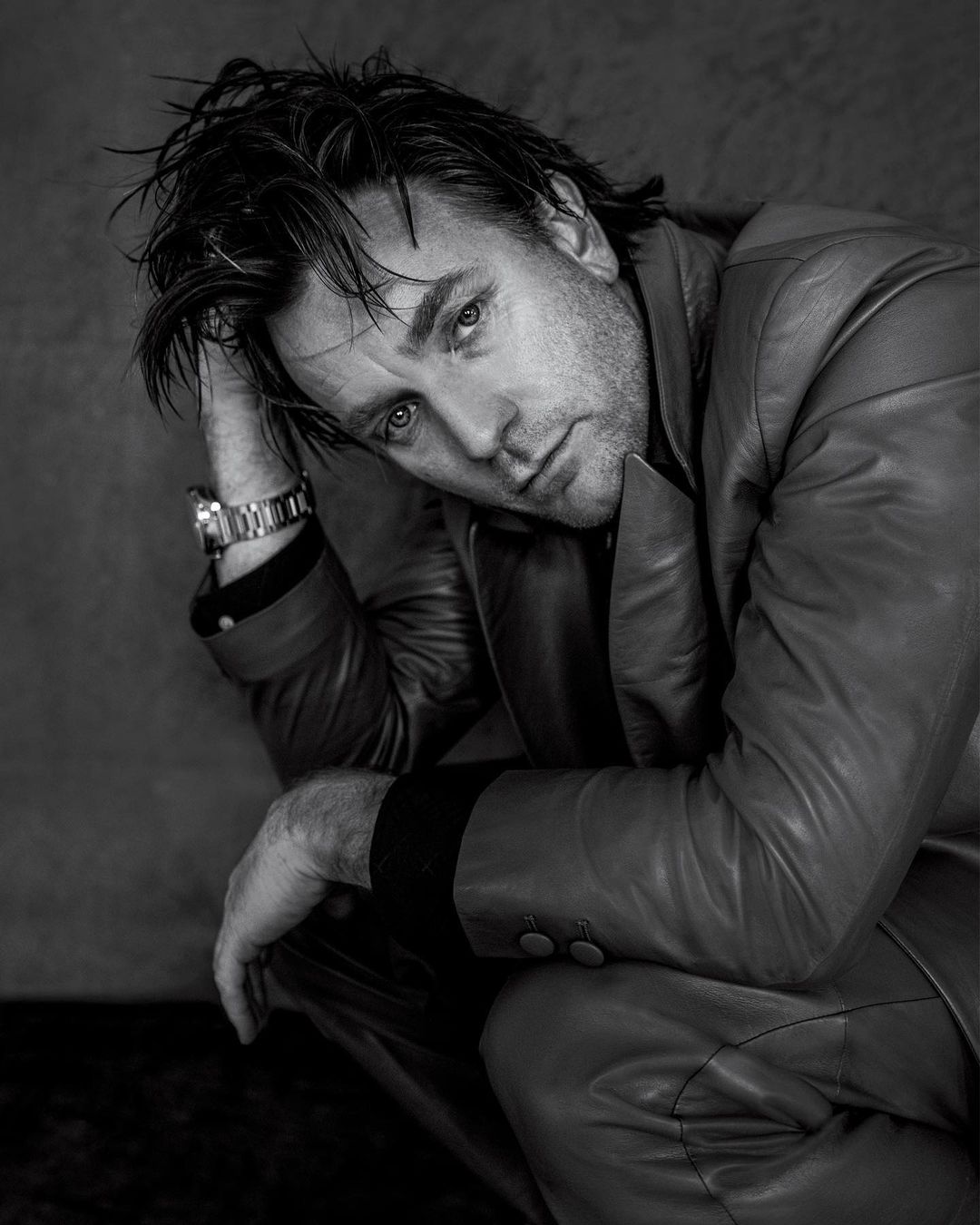 Black and white portrait of a handsome adult man in a leather jacket, seated and looking at the camera with a serious expression.