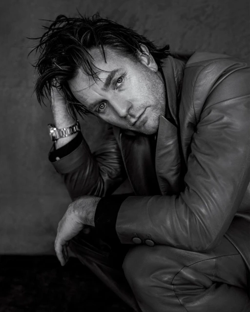 Black and white portrait of a handsome adult man in a leather jacket, seated and looking at the camera with a serious expression.