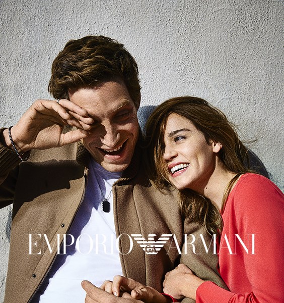 A smiling man and woman leaning against a textured grey wall outdoors in bright sunlight.