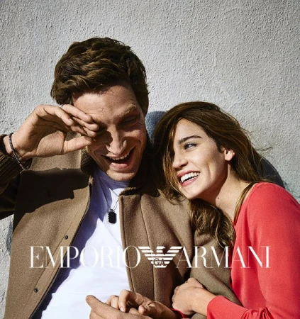 A smiling man and woman leaning against a textured grey wall outdoors in bright sunlight.