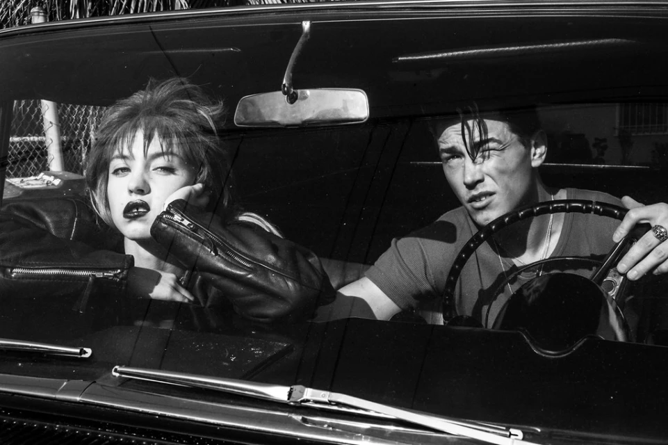 Black and white photograph of a young adult couple in a vintage car. The woman wears a leather jacket, and the man is at the steering wheel.
