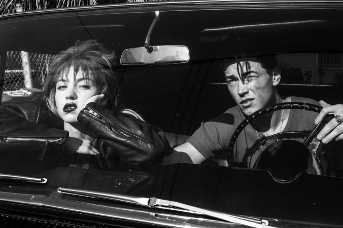 Black and white photograph of a young adult couple in a vintage car. The woman wears a leather jacket, and the man is at the steering wheel.