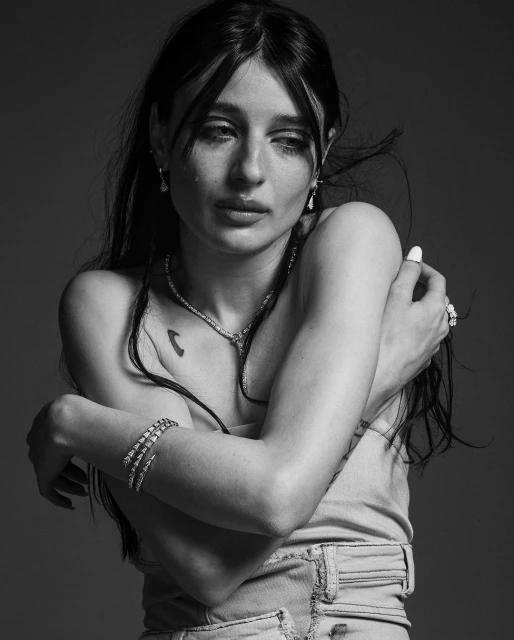 Monochrome portrait of a crying woman with long dark hair, wearing casual clothes and jewelry, with her arms crossed over her chest.