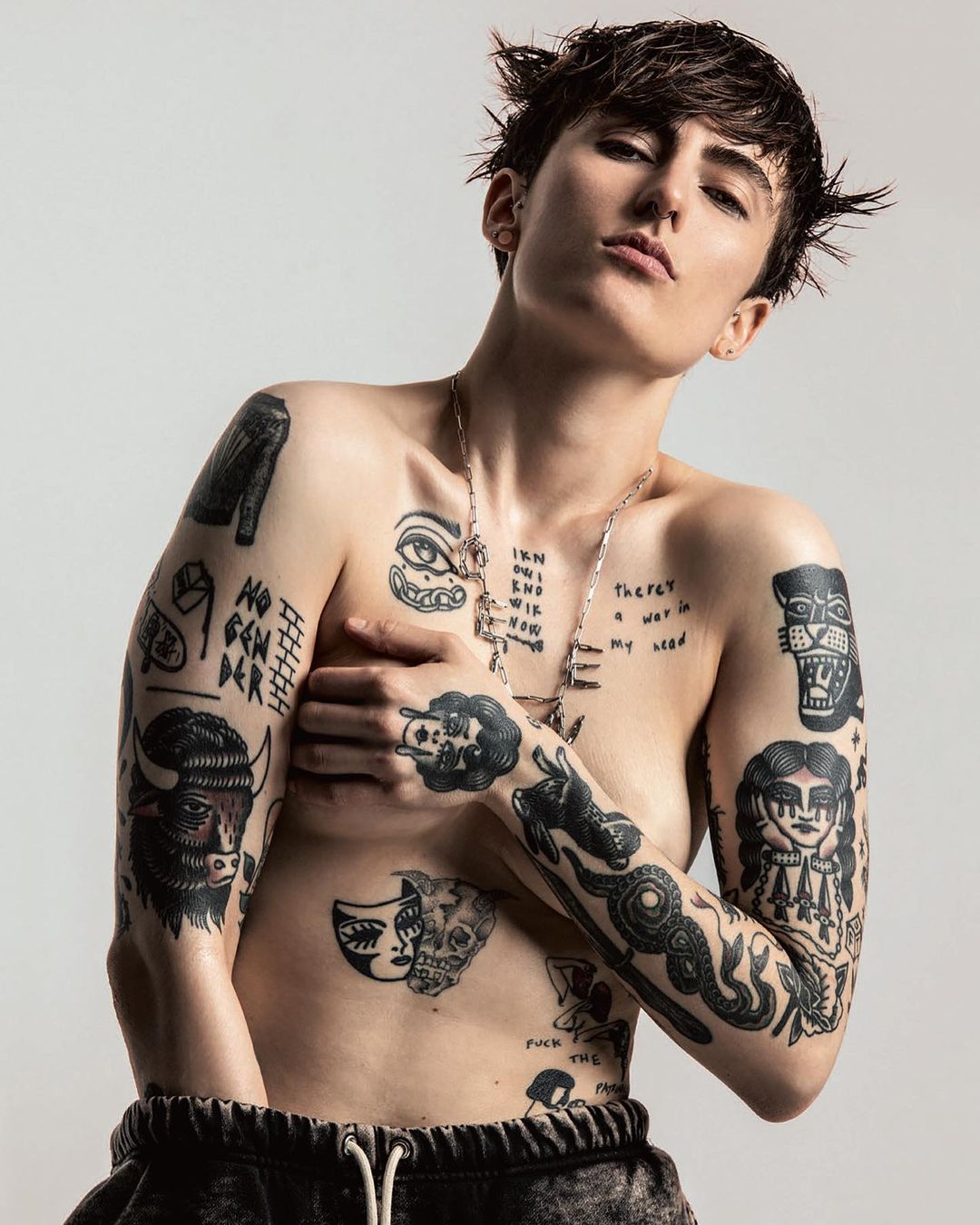 Tattooed person with short hair in a studio portrait, arms partially covering the chest, wearing sweatpants and a chain necklace.