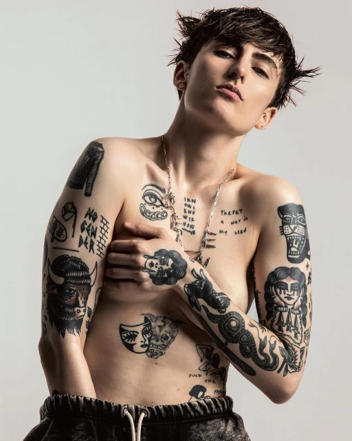 Tattooed person with short hair in a studio portrait, arms partially covering the chest, wearing sweatpants and a chain necklace.