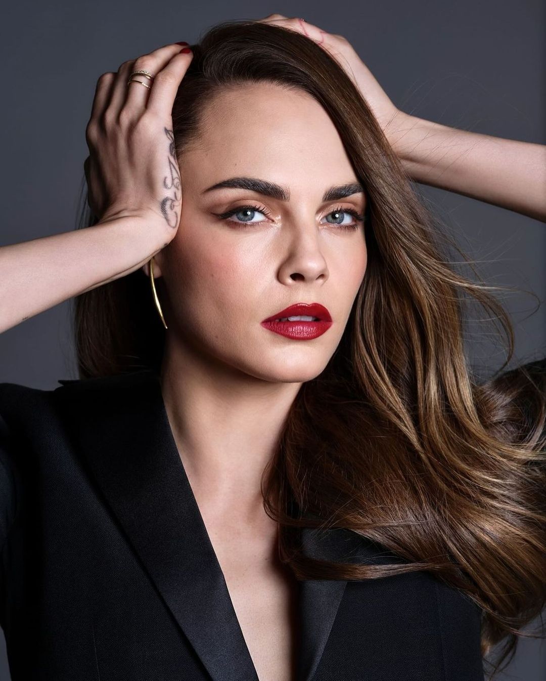 Close-up portrait of Cara Delevingne with hands near her head, wearing a black blazer, with dark red lipstick and light brown hair.