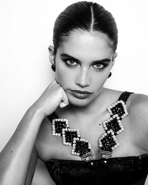 Black-and-white portrait of a woman with slicked-back hair and striking eye makeup, wearing a decorative collar, set against a plain white backdrop.