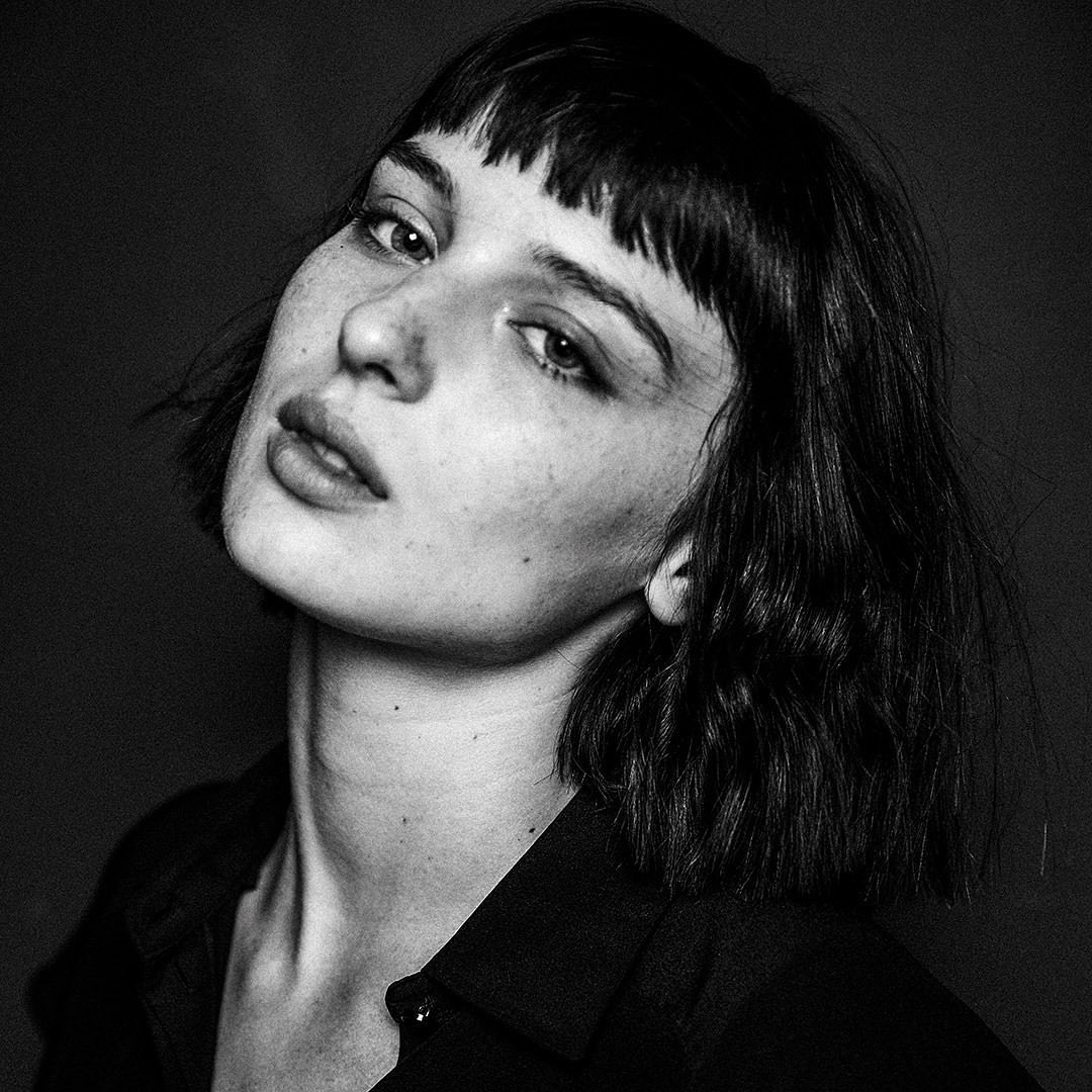 Black and white portrait of a young adult woman with bangs and dark hair, wearing a dark collared shirt against a dark background.