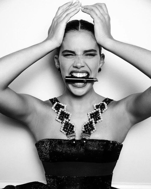 Black and white portrait of a woman with dark hair, wearing a black dress and jeweled necklace, holding a lipstick in her mouth.