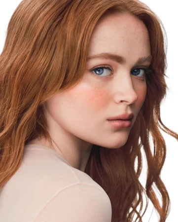 Close-up portrait of a young woman with long red hair and blue eyes looking over her shoulder against a plain white background.