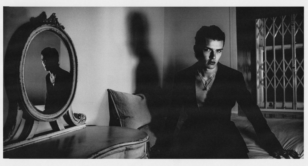 Monochrome portrait of a dark-haired adult male in a blazer leaning against a bed with a vintage mirror and window in the background.