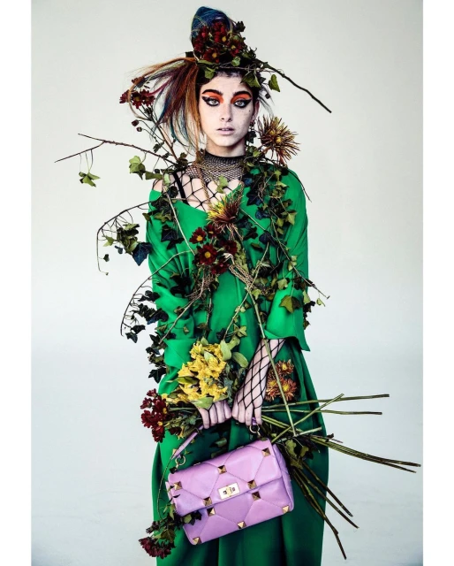 Female model in a green dress and floral headpiece holds a bouquet and pink purse against a white background in a fashion editorial.