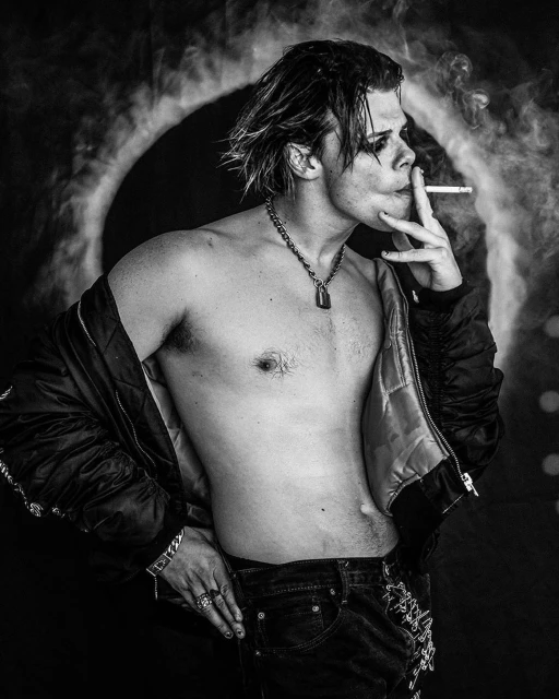 Black and white portrait of a shirtless young adult man smoking a cigarette indoors, wearing dark jeans and jacket.