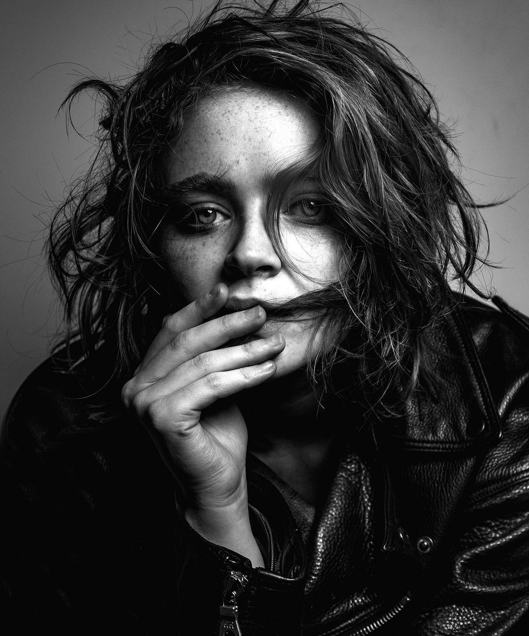 Black and white portrait of a young woman with freckles, wearing a leather jacket, and holding a strand of hair in front of her mouth.