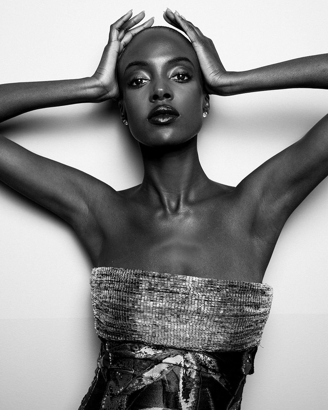 Striking black and white portrait of a dark-skinned adult female model with hands behind her head, wearing a textured strapless top against a plain background.