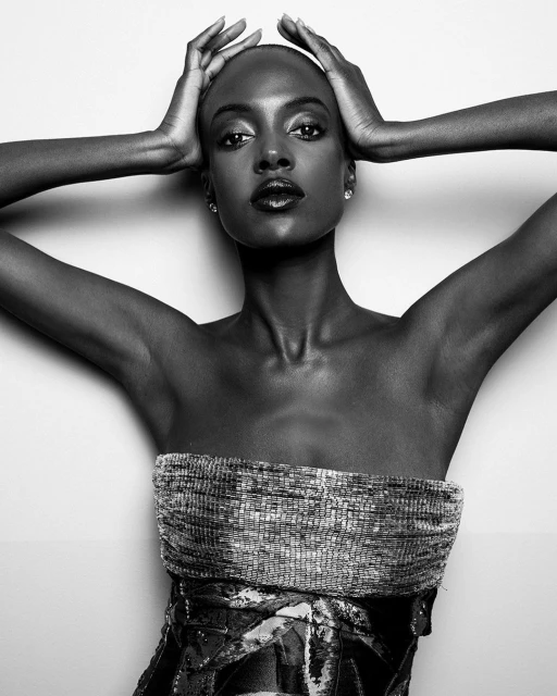 Striking black and white portrait of a dark-skinned adult female model with hands behind her head, wearing a textured strapless top against a plain background.
