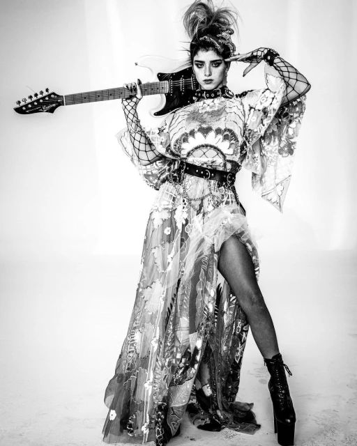 A stylish adult female poses in a flowing dress with floral accents, holding an electric guitar in a black-and-white photo studio setting.