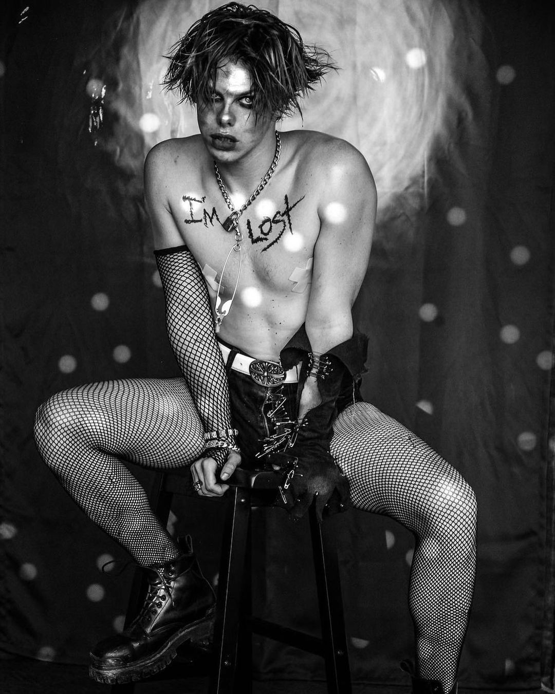 Black and white studio shot of a young adult wearing fishnet stockings and a corset, sitting on a stool with text on torso.