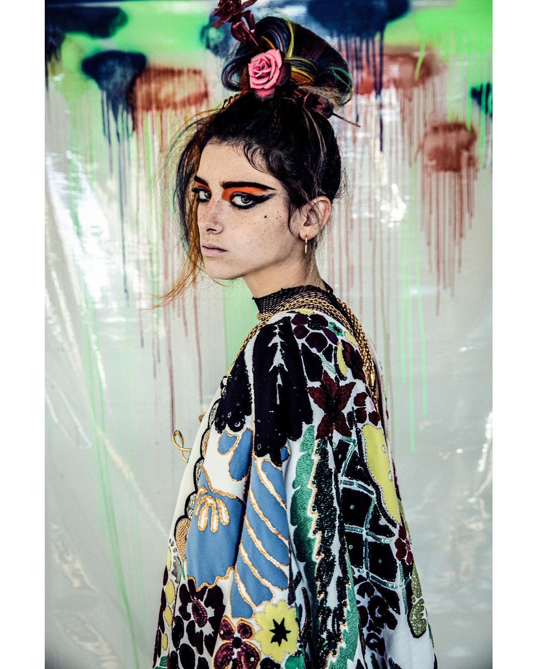 Portrait of a stylish young woman with eye makeup and a flower hair accessory in a colorful patterned jacket, posing against a vibrant backdrop.