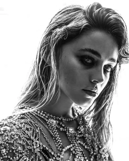 Monochrome portrait of a young woman with wet blonde hair, wearing ornate jeweled top and makeup in a bright white indoor setting.