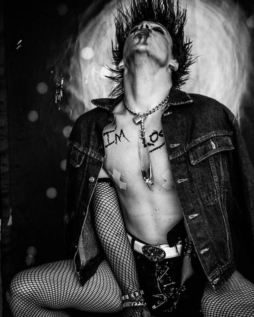 A person with a mohawk and denim jacket looks upward in a black and white image with fishnet stockings and visible chest tattoos.