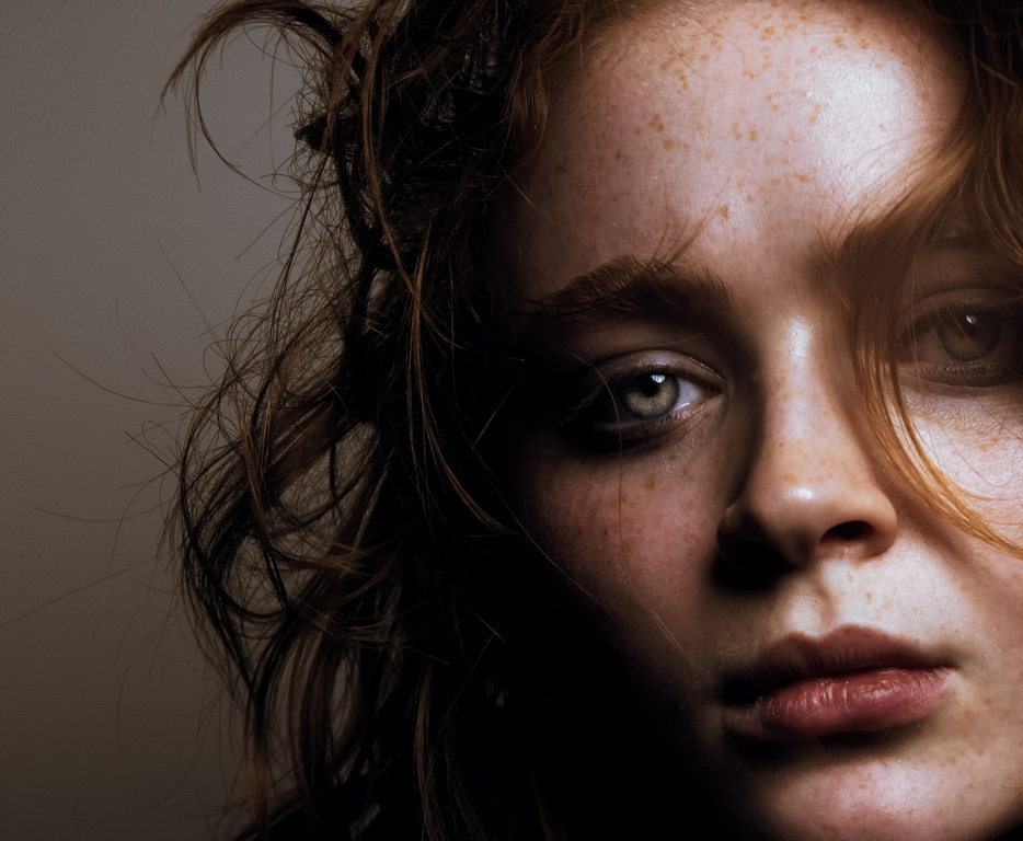 Close-up portrait of a fair-skinned adult female with freckles, looking directly at the camera in diffused, soft lighting conditions.