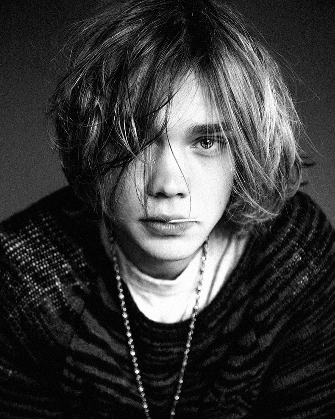 Monochrome portrait of a young adult with shoulder-length hair, wearing a patterned sweater and a beaded necklace. Close-up studio shot.