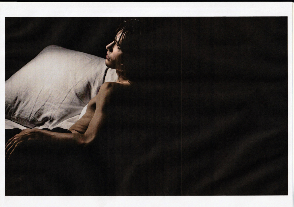 Man lying in bed on white pillow in dark, intimate setting. His skin contrasts with the dark background. He gazes off to the side.