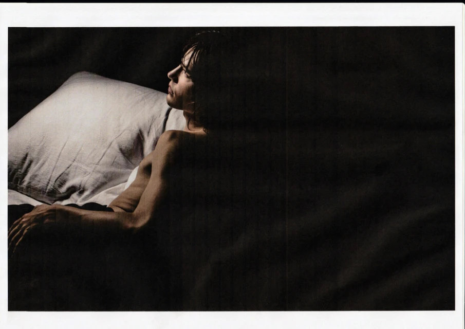 Man lying in bed on white pillow in dark, intimate setting. His skin contrasts with the dark background. He gazes off to the side.