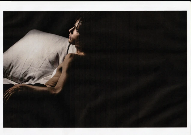 Man lying in bed on white pillow in dark, intimate setting. His skin contrasts with the dark background. He gazes off to the side.