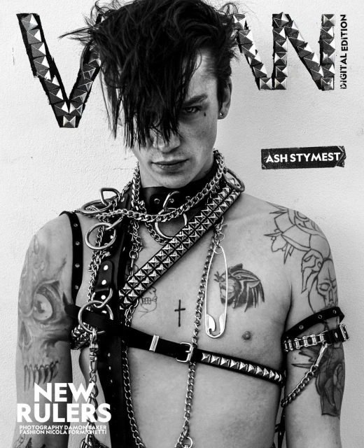 Monochrome portrait of Ash Stymest on a magazine cover, wearing leather harness and chains. The background is off-white, and the image features tattoos.