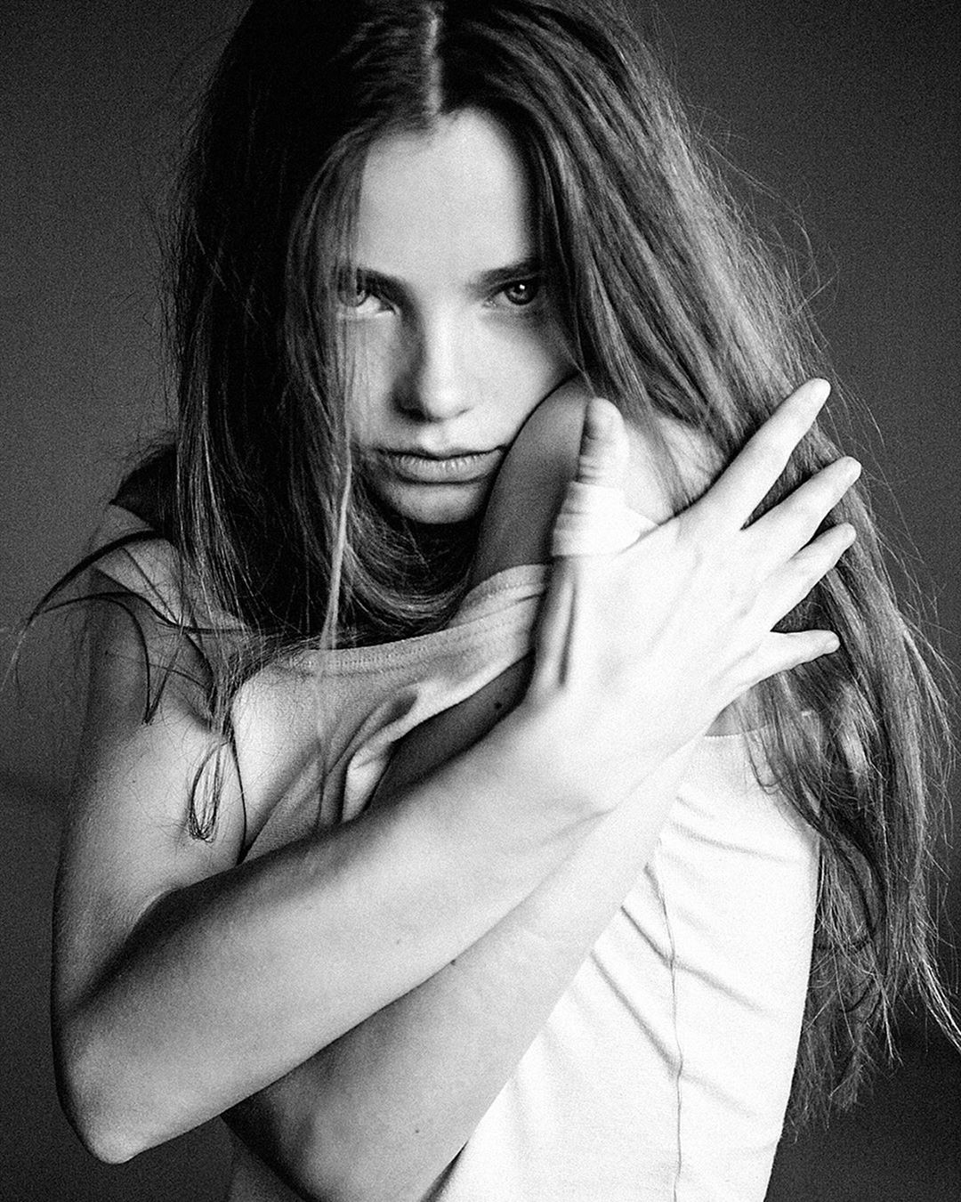 Black and white portrait of a young adult woman with long hair, embracing herself in a photo studio setting with diffused light.
