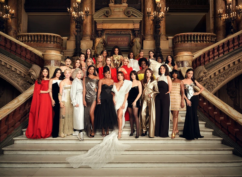 Large group of diverse women posing on a grand staircase in formal dresses, inside an ornate building with high ceilings and chandeliers.