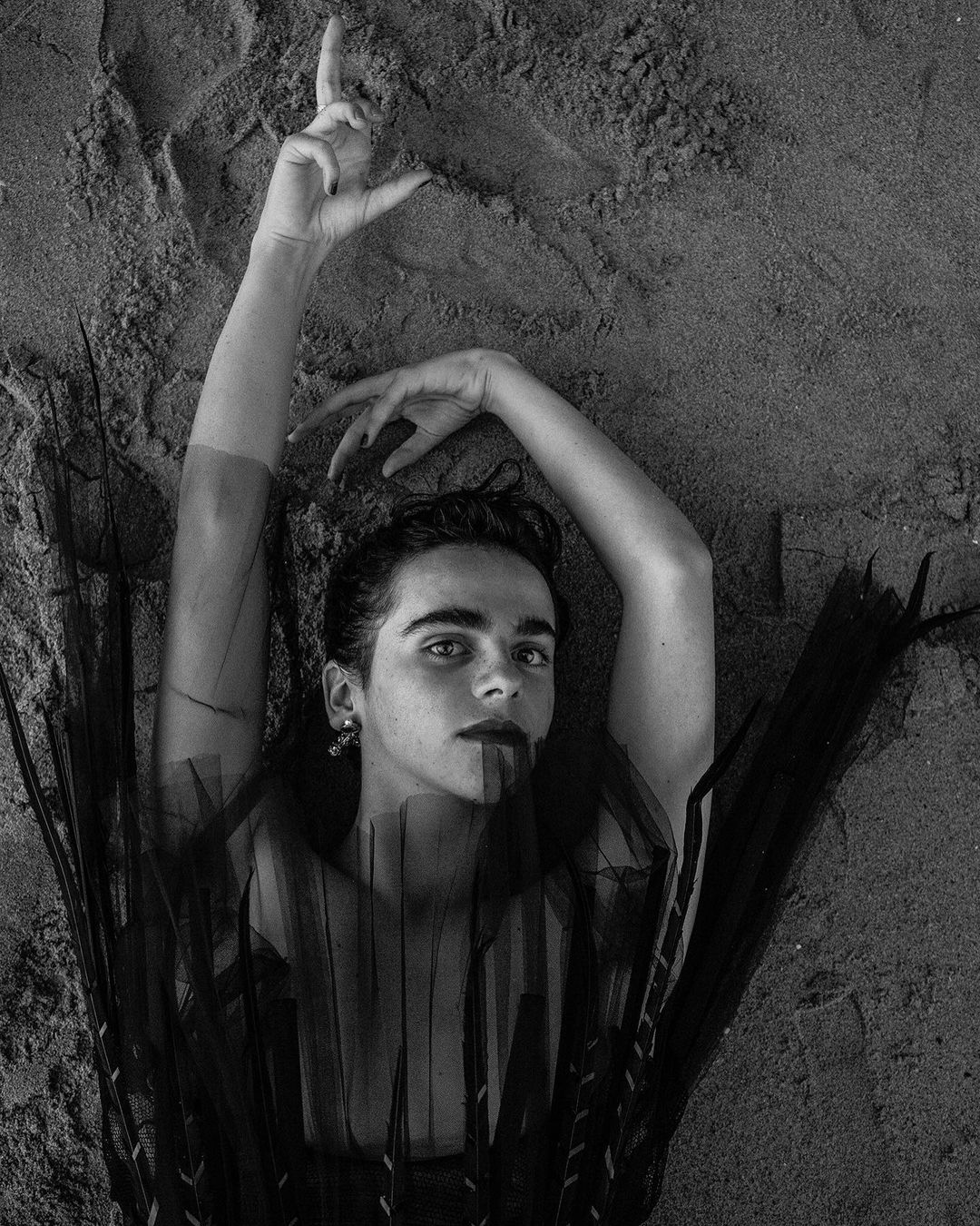 Monochrome portrait of a young adult laying on sand, adorned with tulle and dark makeup, arms raised in expressive pose. 