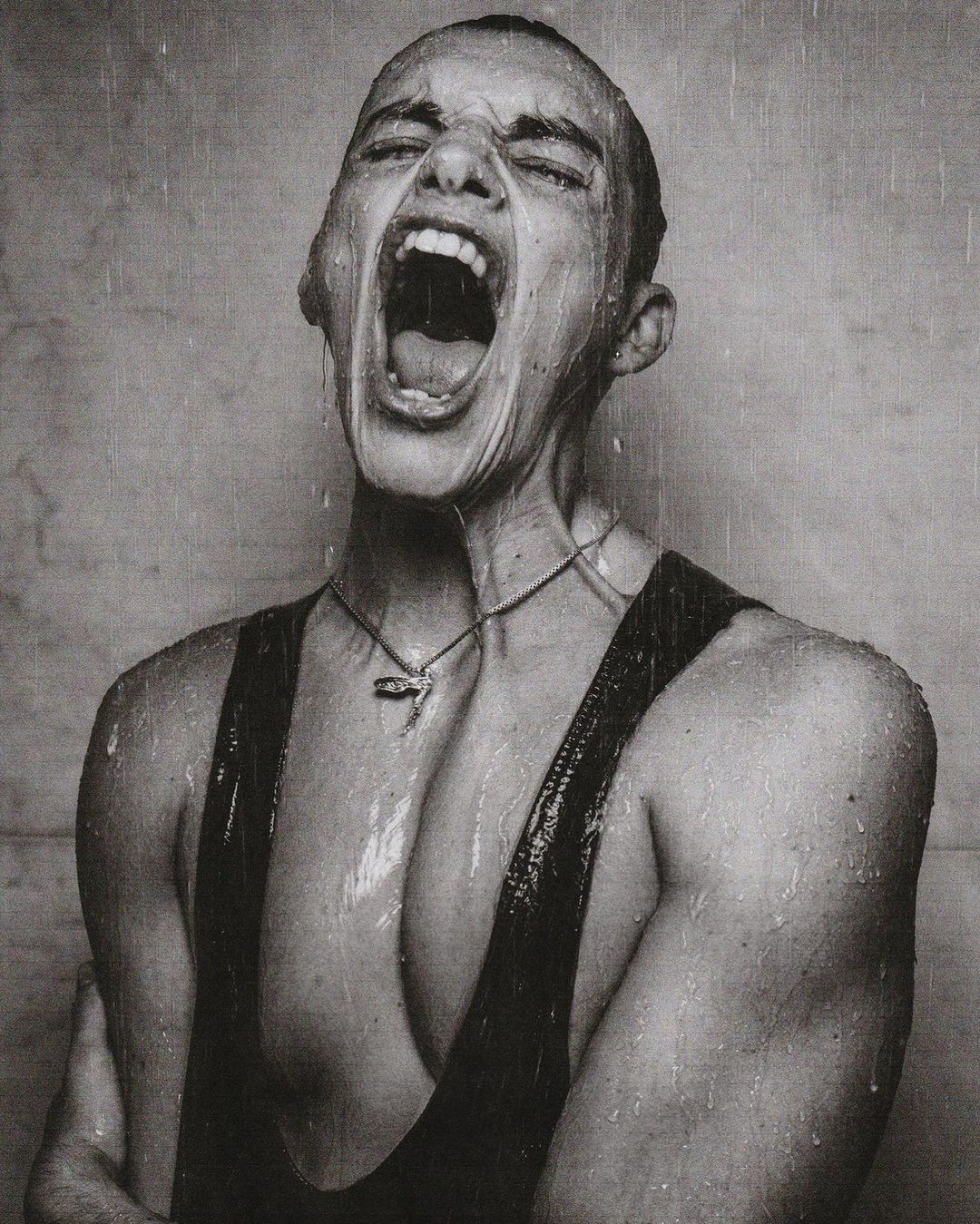Monochrome portrait of a young adult with short hair, mouth wide open screaming in the rain, wearing a tank top and necklace.