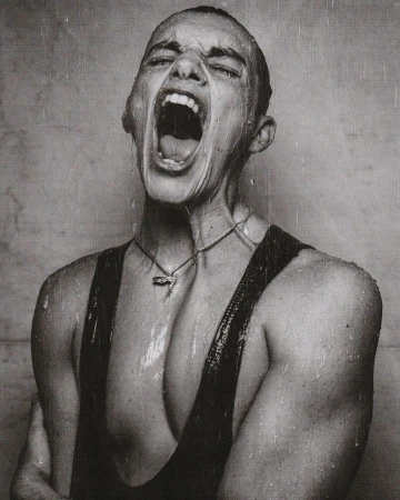 Monochrome portrait of a young adult with short hair, mouth wide open screaming in the rain, wearing a tank top and necklace.