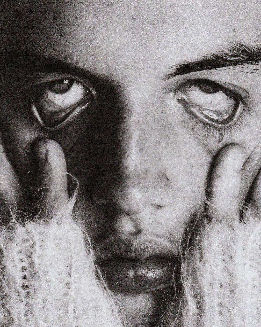 Close-up monochrome portrait of a person with hands near the face, looking upwards, wearing a textured sweater, conveying a fine-art aesthetic.