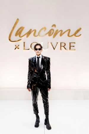 A person in a black patent leather suit and boots stands against a Lancôme x Louvre branded backdrop in a fashion-forward presentation.